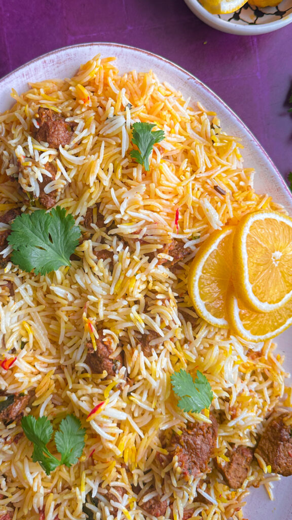 saffron rice mixed with lamb, garnished with cilantro and lemon slices plated on an oval dish on a deep purple background
