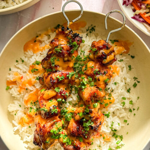 Bowl of cooked skewered chicken over white rice, drizzled with orange bang bang sauce garnished with parsley and chives