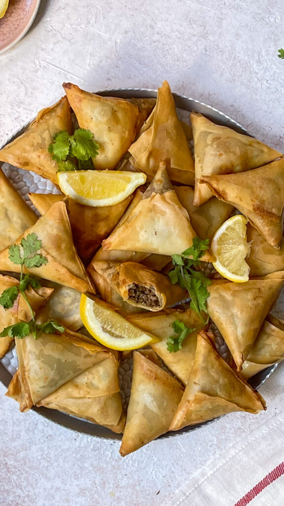 plate of crispy samosas with lemon slices and cilantro to garnish. one samosa bit open to show ground beef filling